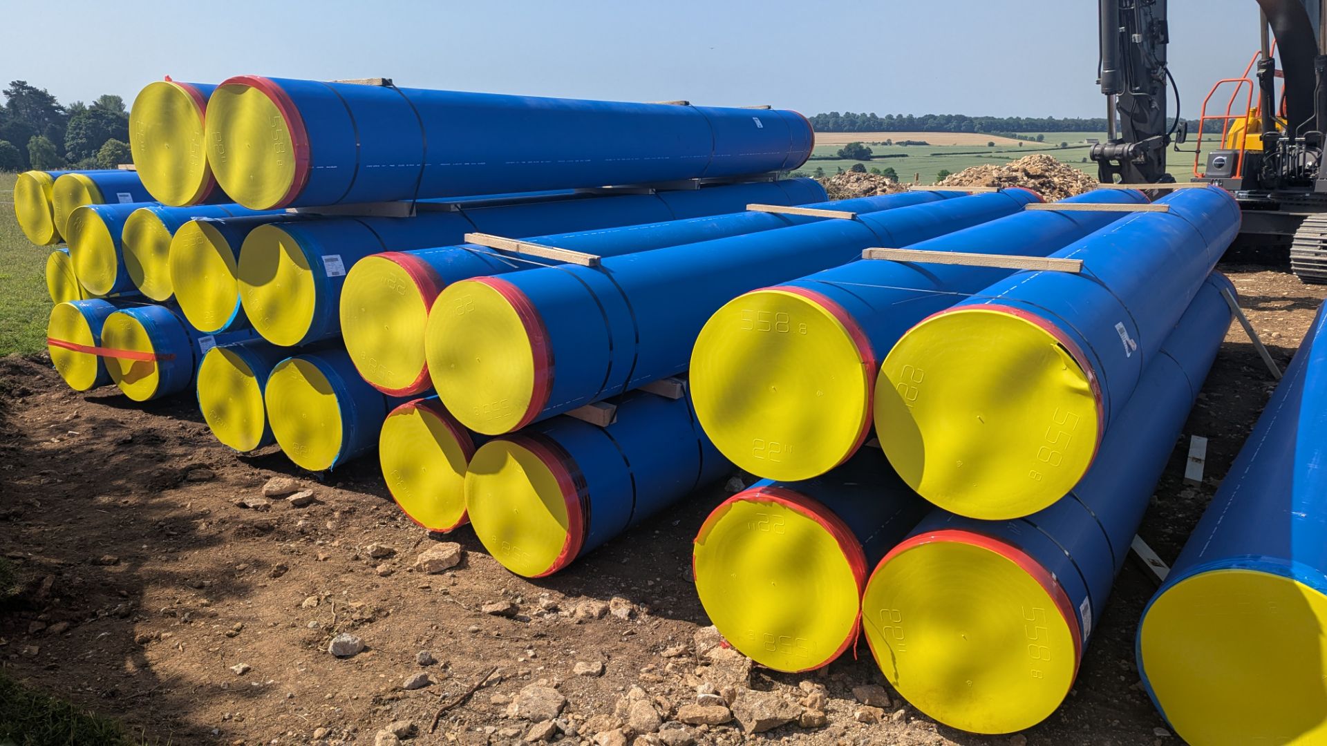Stacks of large blue PE water pipe with yellow end caps on a pipeline site, with an excavator behind